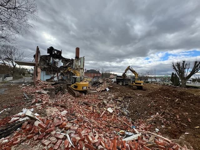 Two excavators demolishing a brick building with a dump truck standing by